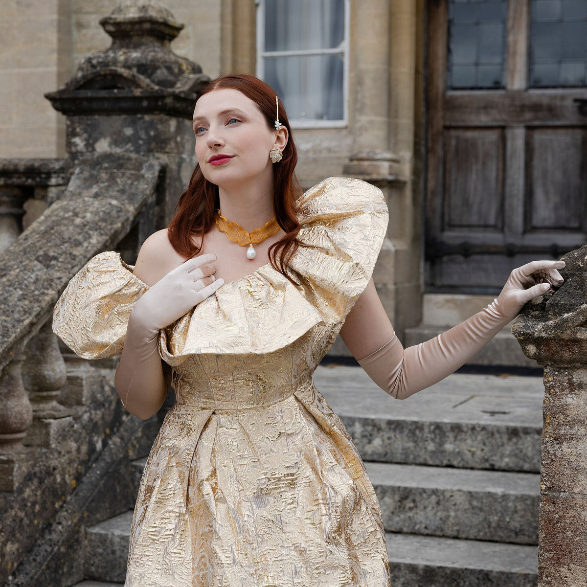 Woman wearing long, below elbow satin gloves in champagne beige in a shiny gold dress on stone steps.