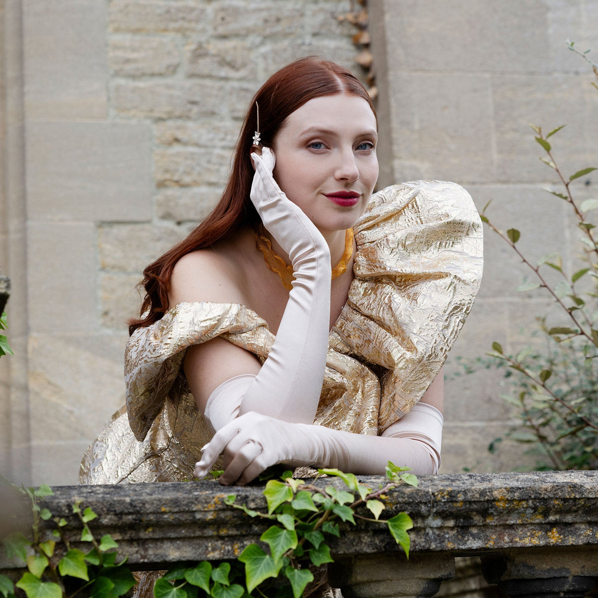 Woman wearing long, below elbow satin gloves in champagne beige in a shiny gold dress leaning on balcony stone.