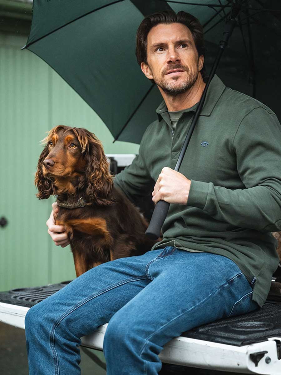Man sitting on a vehicle's tailgate with a dog, holding an umbrella.