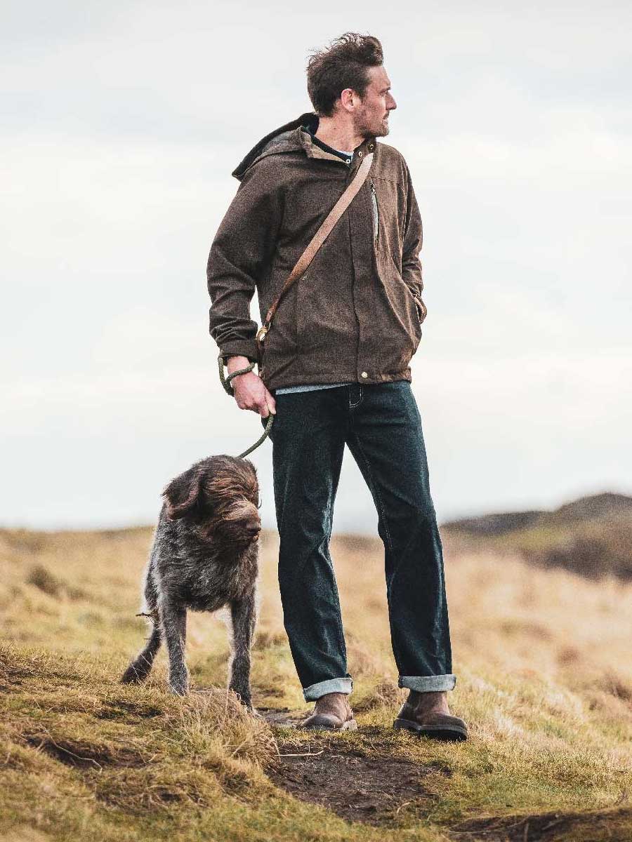 Man walking a dog in a field wearing a brown jacket and dark jeans.