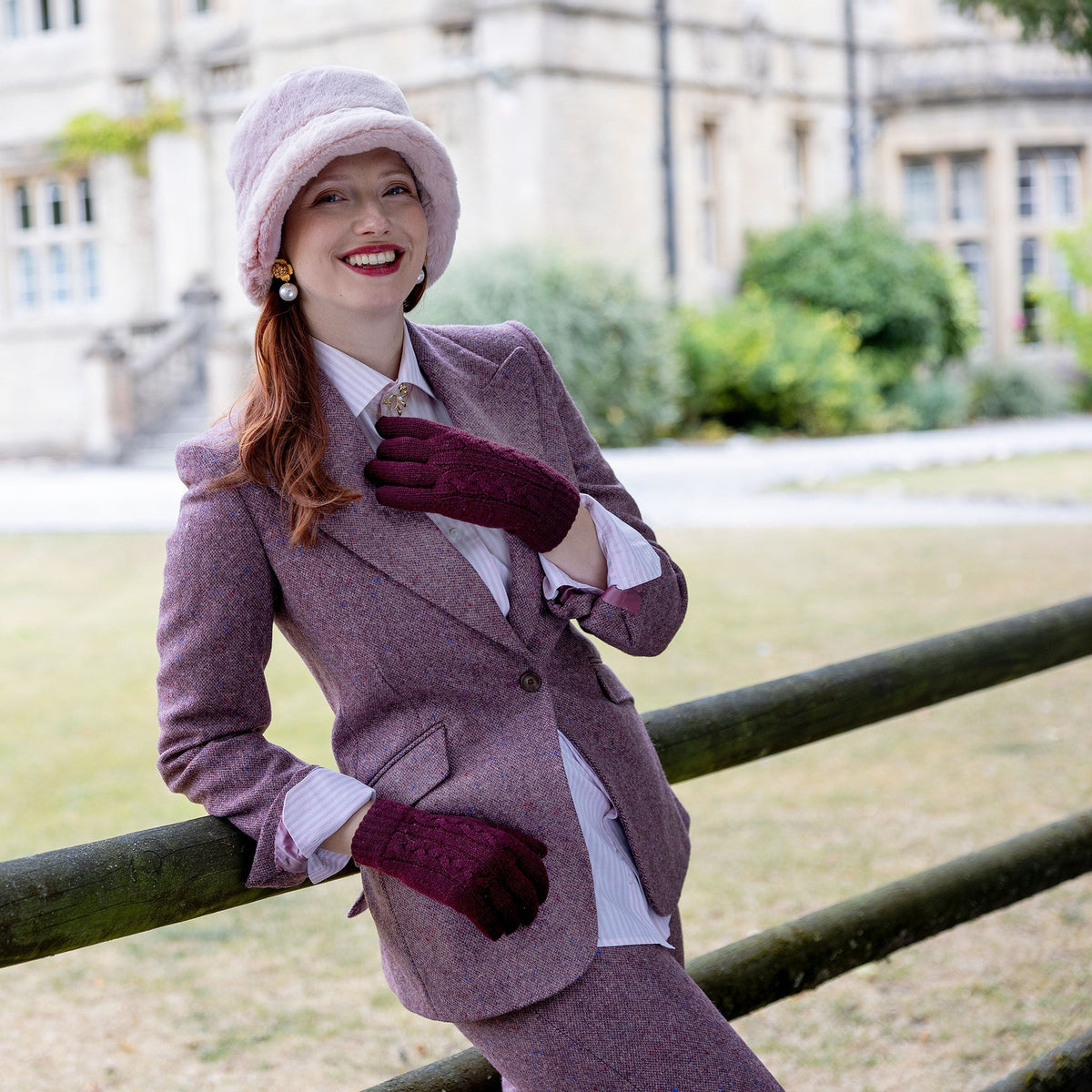 Woman wearing cable knit gloves in burgundy, in a pink suit leaning against a fence. 