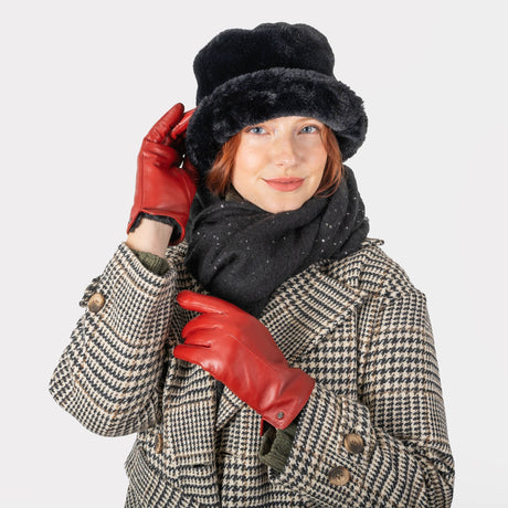 Woman wearing touchscreen single point faux fur lined leather gloves in red, in a check coat and black hat and scarf. With white background.