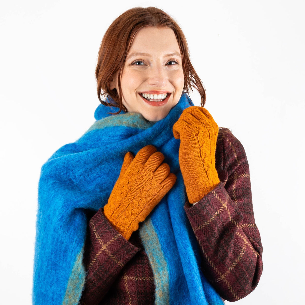Woman wearing cable knit gloves in pumpkin with a blue scarf and burgundy coat with white background. 