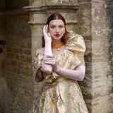 Woman wearing light pink satin below elbow gloves in gold shiny dress in a stone archway. 