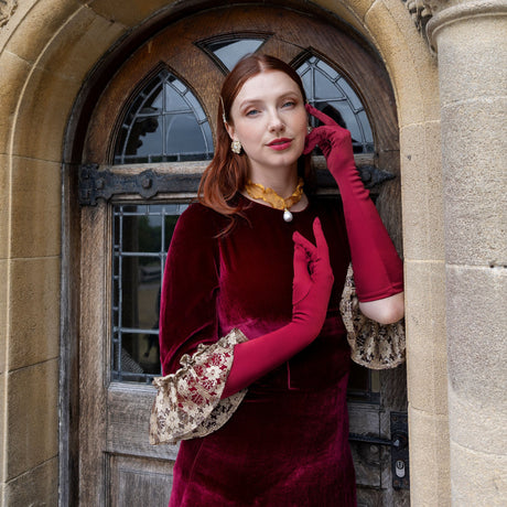 Woman wearing burgundy satin below elbow gloves in burgundy dress in a doorway. 