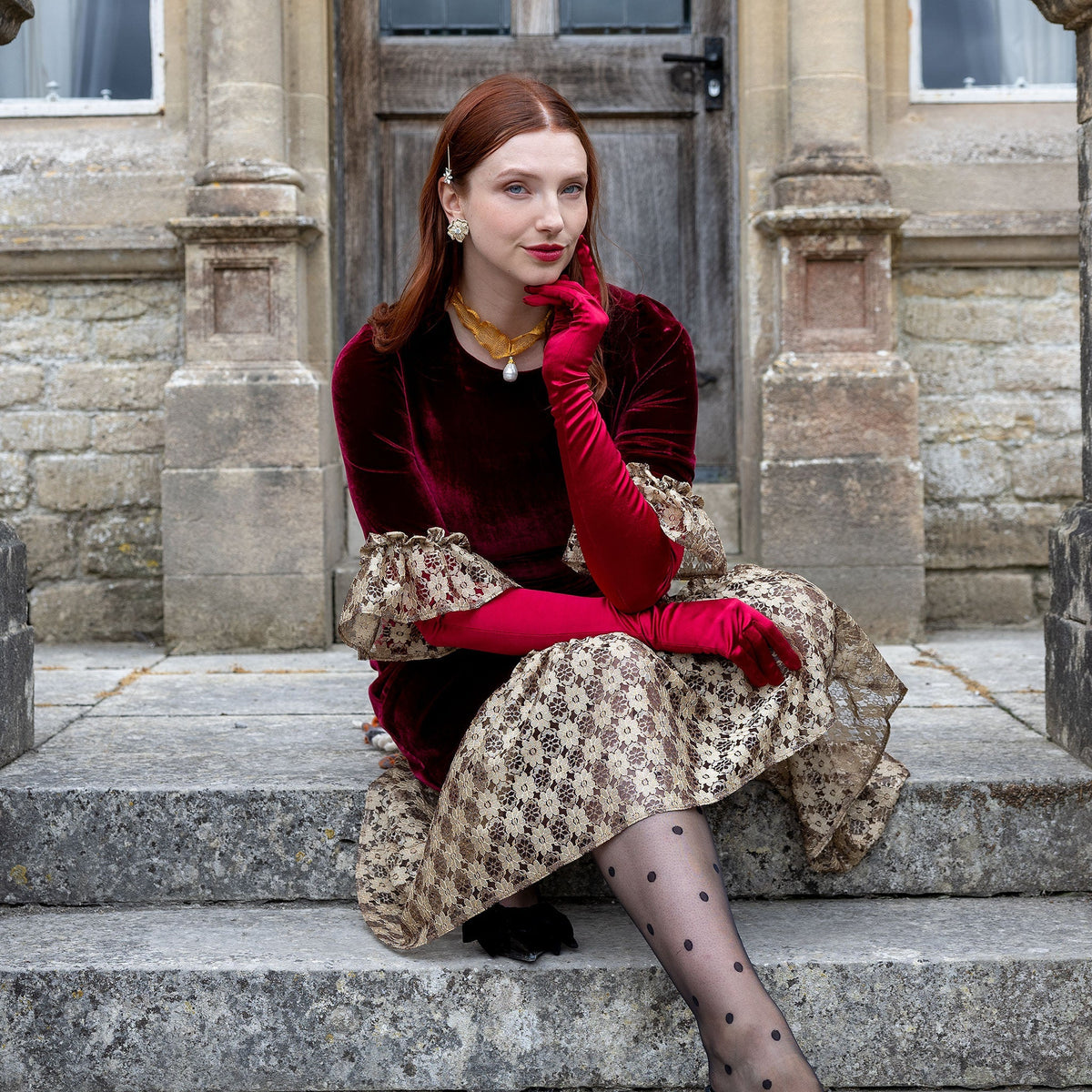 Woman wearing burgundy satin below elbow gloves in burgundy dress on stone steps. 