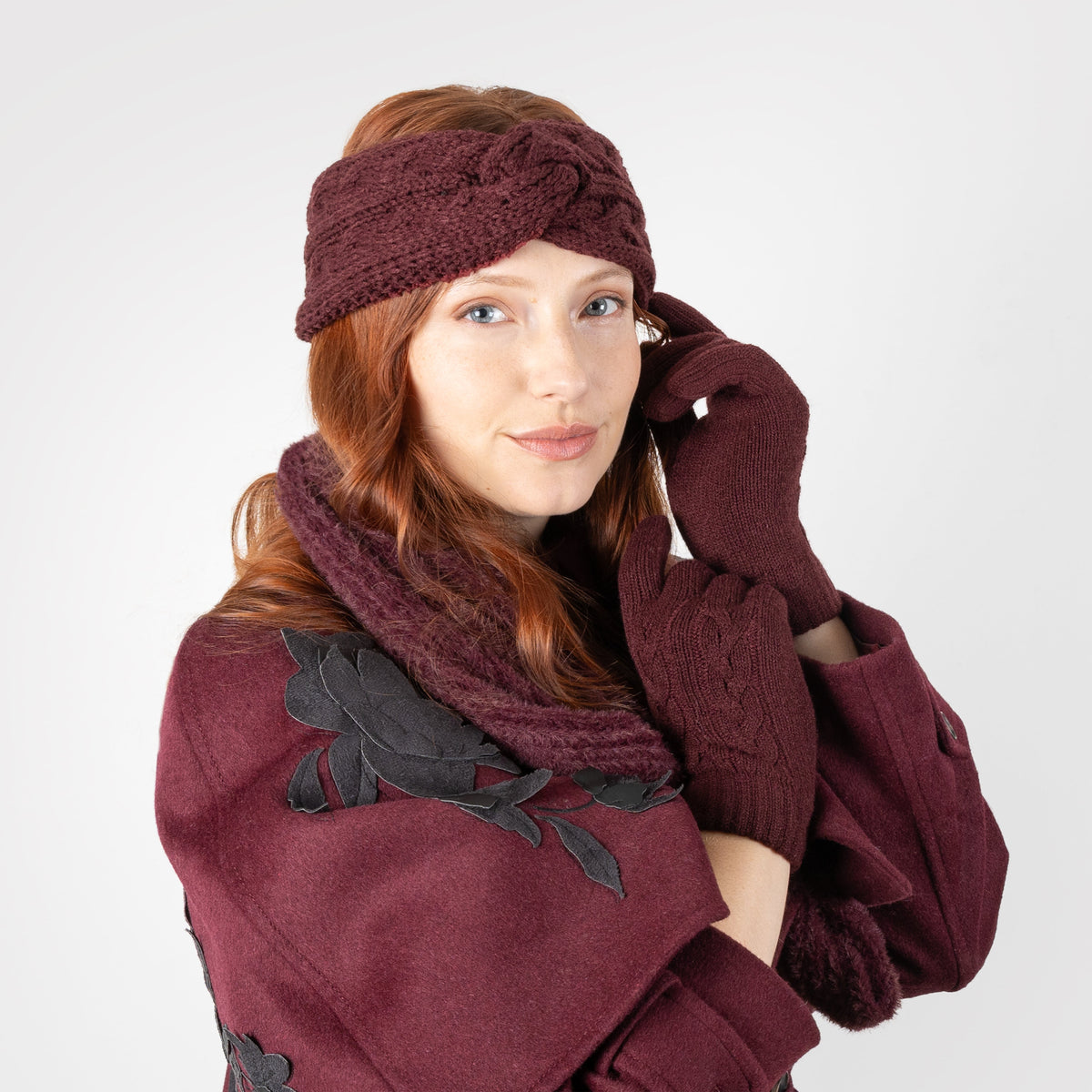 Woman wearing cable knit gloves in burgundy, in burgundy coat and matching scarf and headband. 