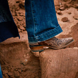Person wearing blue jeans and brown cowboy boots stepping onto a rock in a natural setting.