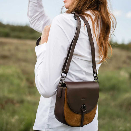 A woman in a white shirt carries a brown leather and suede crossbody bag outdoors in a grassy field. The bag has a dark brown flap, a tan suede lower section, and a dark brown shoulder strap.