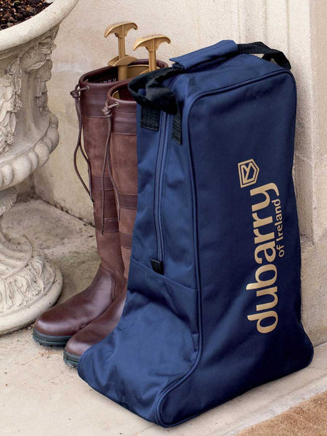 A navy blue Dubarry of Ireland boot bag with gold lettering, standing on a stone ledge next to a pair of brown leather riding boots on wooden shoe trees.