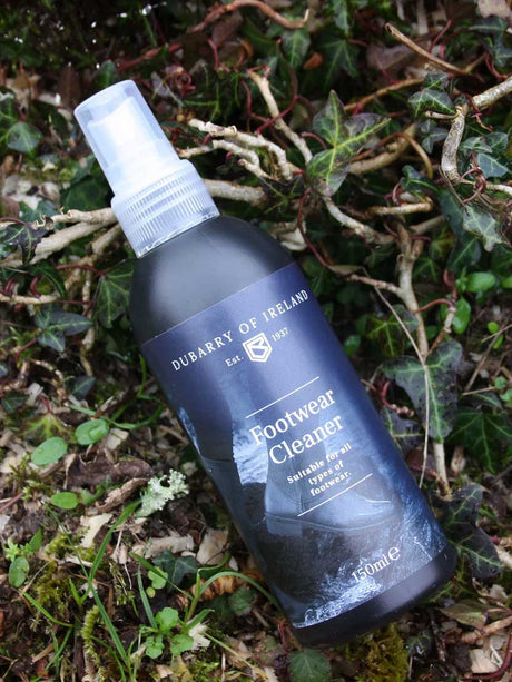 A bottle of Dubarry of Ireland Footwear Cleaner lies on a bed of green ivy and dry twigs outdoors. The bottle is a dark charcoal gray with a clear spray nozzle and a navy blue label.