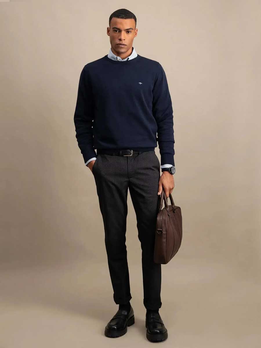 A man wearing a navy blue crewneck sweater over a light blue collared shirt, paired with dark gray trousers, black leather shoes, and holding a brown leather briefcase against a solid beige studio background.