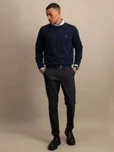 A man wearing a navy blue crewneck sweater over a light blue collared shirt, paired with dark gray trousers and black leather shoes, standing against a neutral beige studio background.