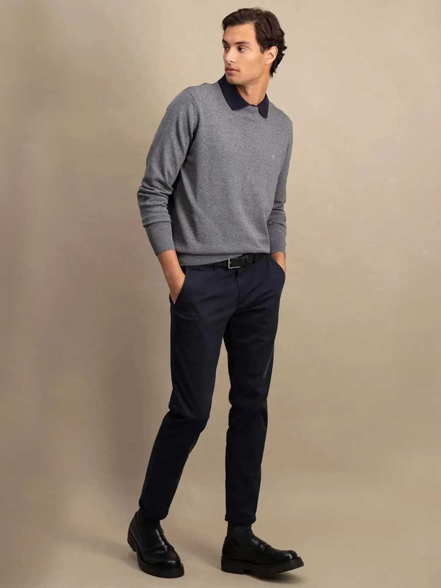 A man modeling a gray long-sleeve polo sweater with a dark navy collar, paired with navy trousers and black leather shoes, against a solid beige studio background.