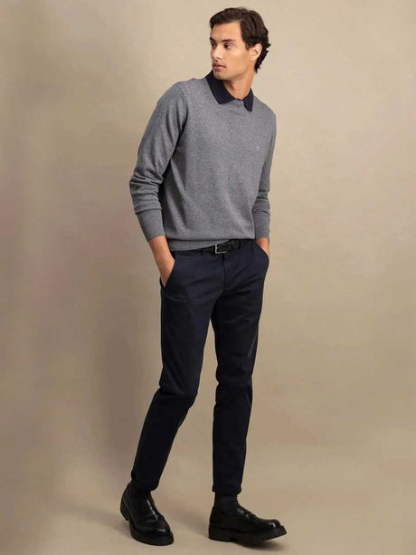 A man modeling a gray long-sleeve polo sweater with a dark navy collar, paired with navy trousers and black leather shoes, against a solid beige studio background.