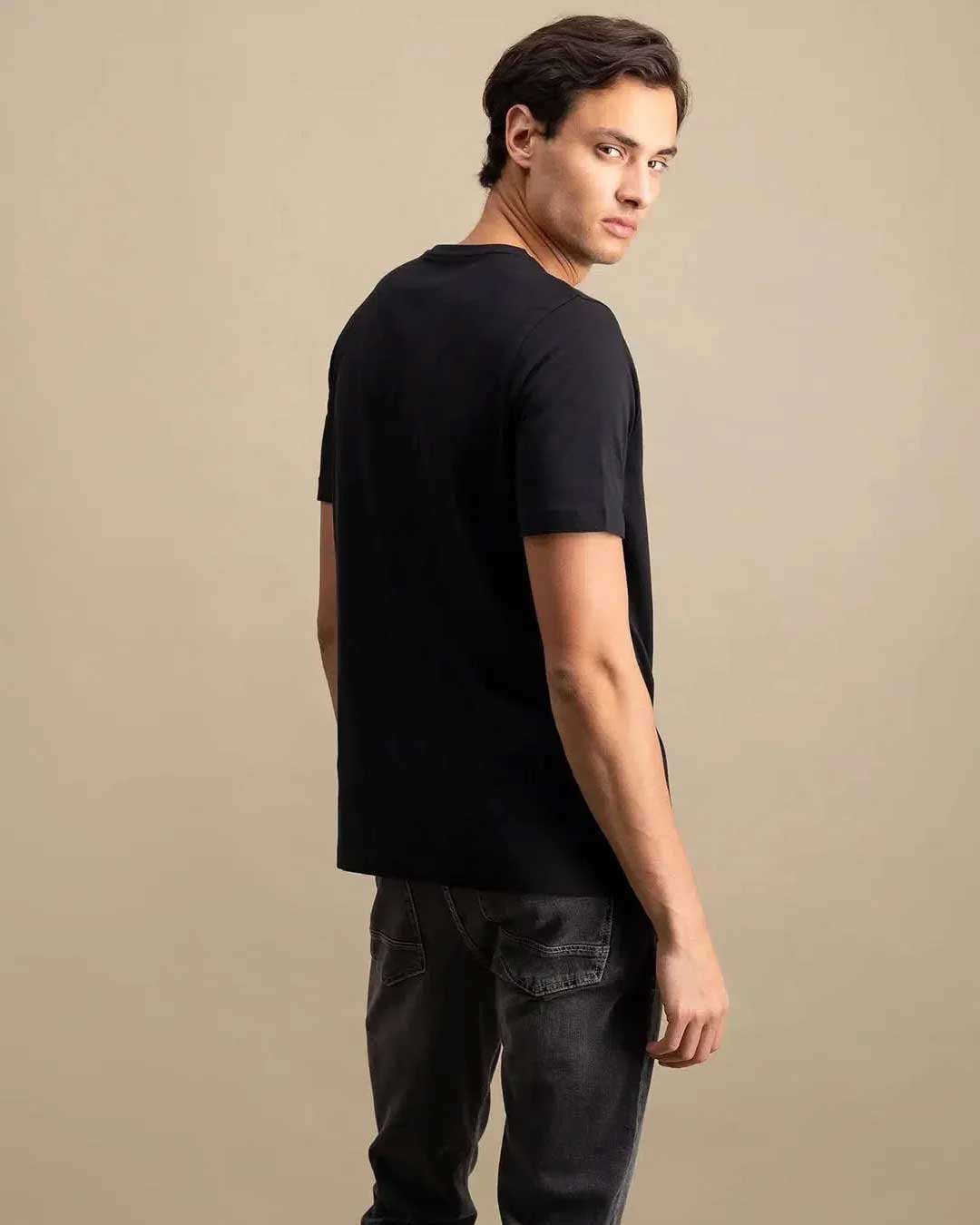 A man wearing a black short-sleeve crewneck t-shirt and dark gray jeans, photographed from a three-quarters back angle against a solid beige studio background.