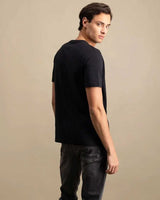 A man wearing a black short-sleeve crewneck t-shirt and dark gray jeans, photographed from a three-quarters back angle against a solid beige studio background.