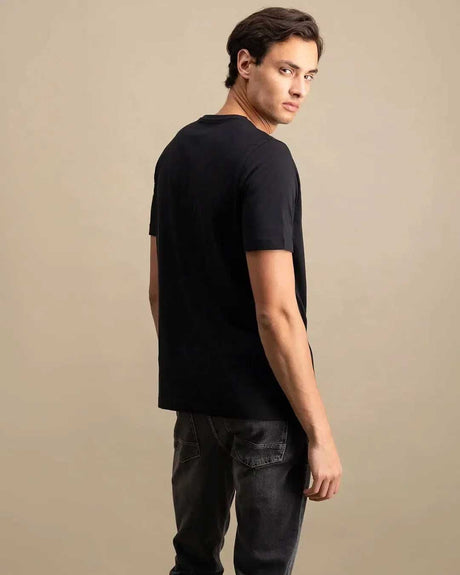A man wearing a black short-sleeve crewneck t-shirt and dark gray jeans, photographed from a three-quarters back angle against a solid beige studio background.