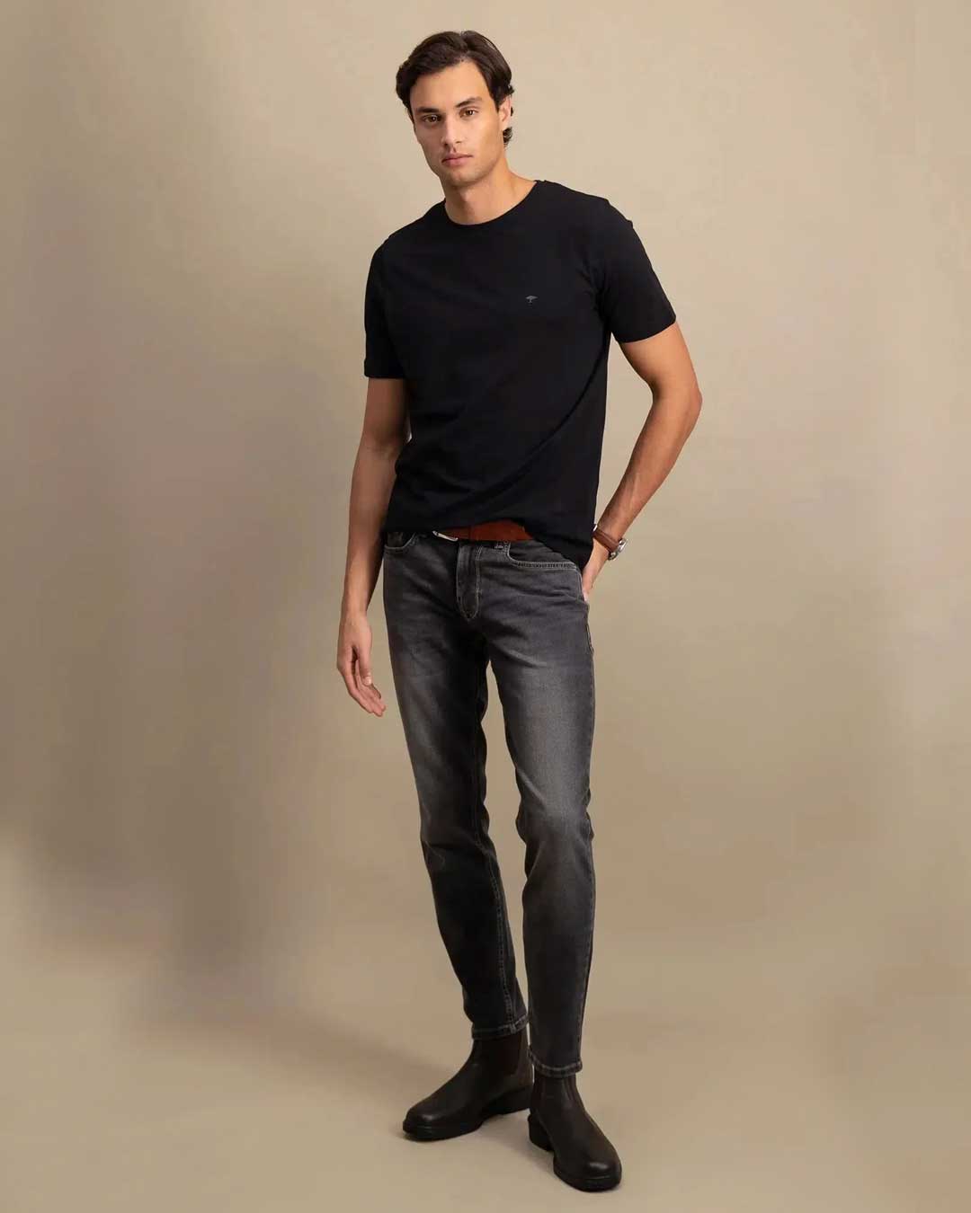 A man modeling a black short-sleeve crewneck t-shirt, dark gray slim-fit jeans, and black leather ankle boots, standing against a solid beige studio background.