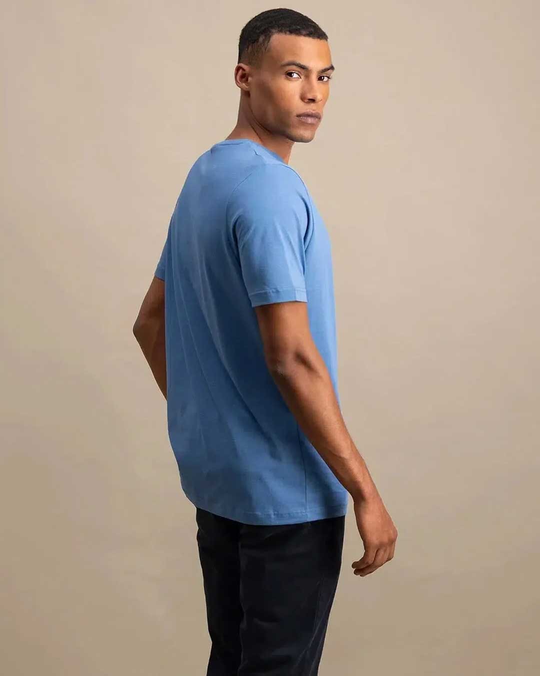 A man wearing a light blue short-sleeve crewneck t-shirt and dark pants, photographed from the side against a plain beige studio background.