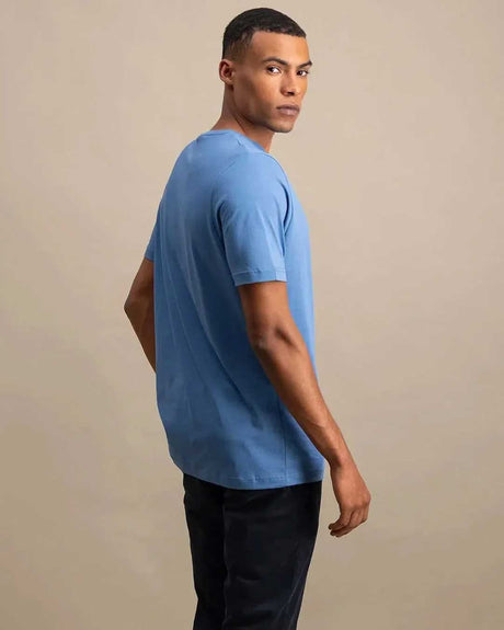 A man wearing a light blue short-sleeve crewneck t-shirt and dark pants, photographed from the side against a plain beige studio background.
