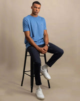 A man wearing a light blue crew-neck t-shirt, dark navy trousers, white socks, and white sneakers sits on a black metal stool against a solid beige studio background.
