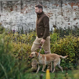 A man in a brown fleece jacket and khaki pants walks a tan dog on a leash through a grassy garden with a white brick wall in the background.