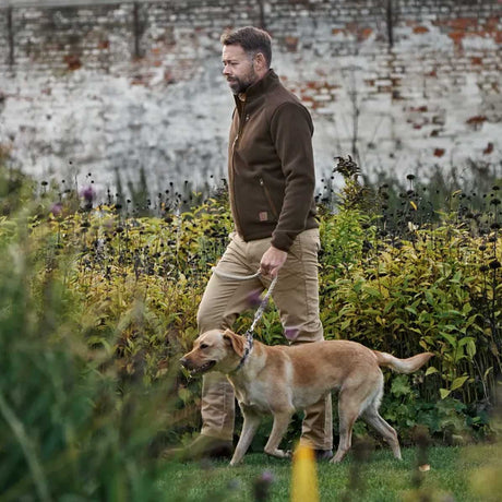 A man in a brown fleece jacket and khaki pants walks a tan dog on a leash through a grassy garden with a white brick wall in the background.