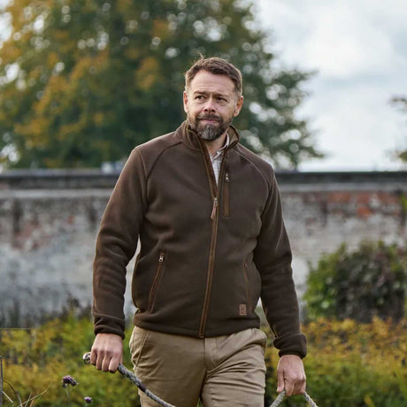 A man wearing a dark brown fleece zip-up jacket and khaki trousers stands outdoors in a garden setting, holding a garden tool.