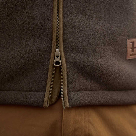 Close-up of a dark brown fleece jacket with a central metal zipper, a brown leather brand patch on the right chest, and a brown garment underneath.