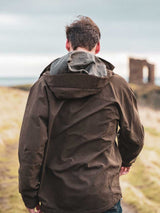 Person wearing a brown jacket standing in a scenic outdoor setting with ruins in the background.