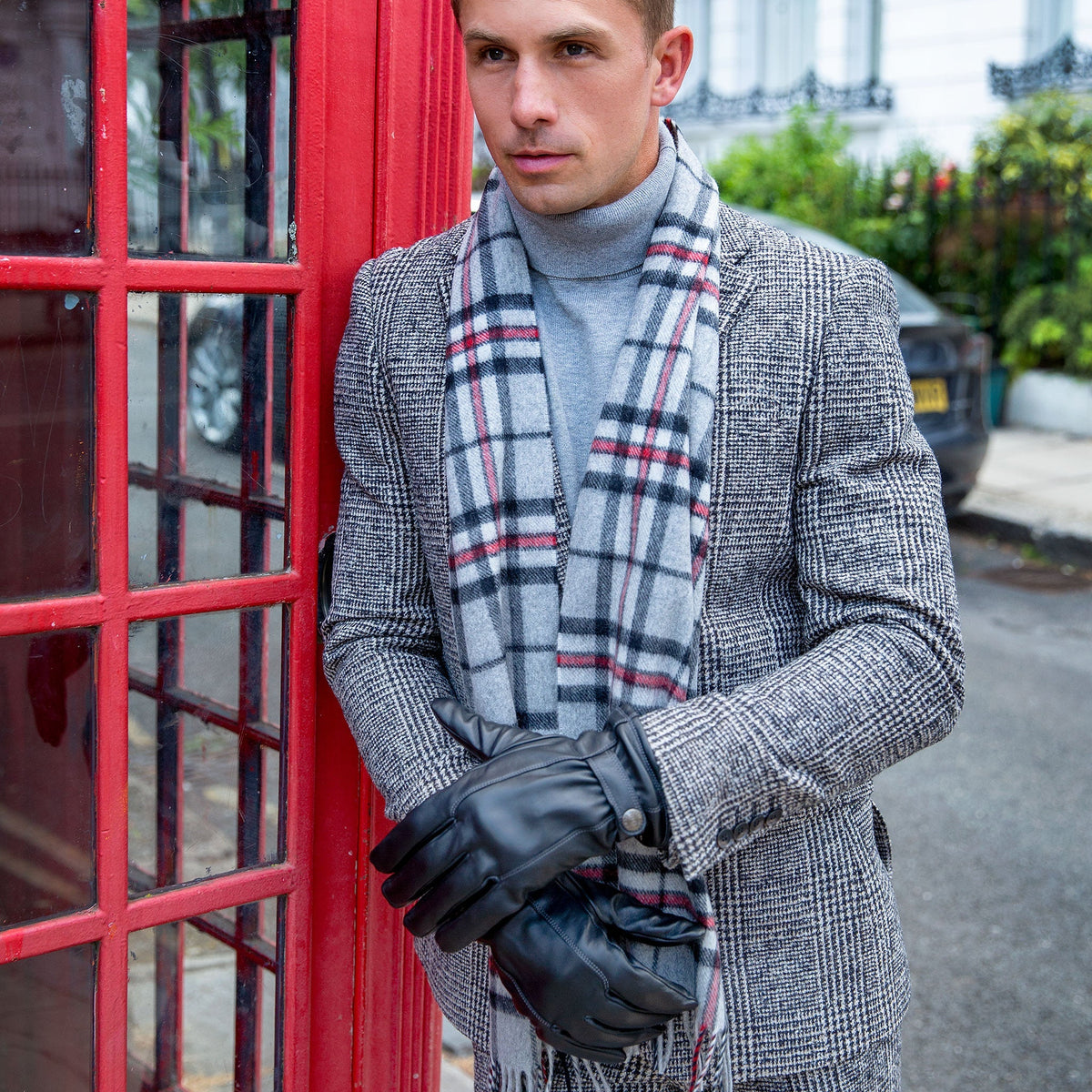 Man wearing touchscreen leather gloves with silver cashmere scarf and tweed jacket leaning on a red phonebooth in the city