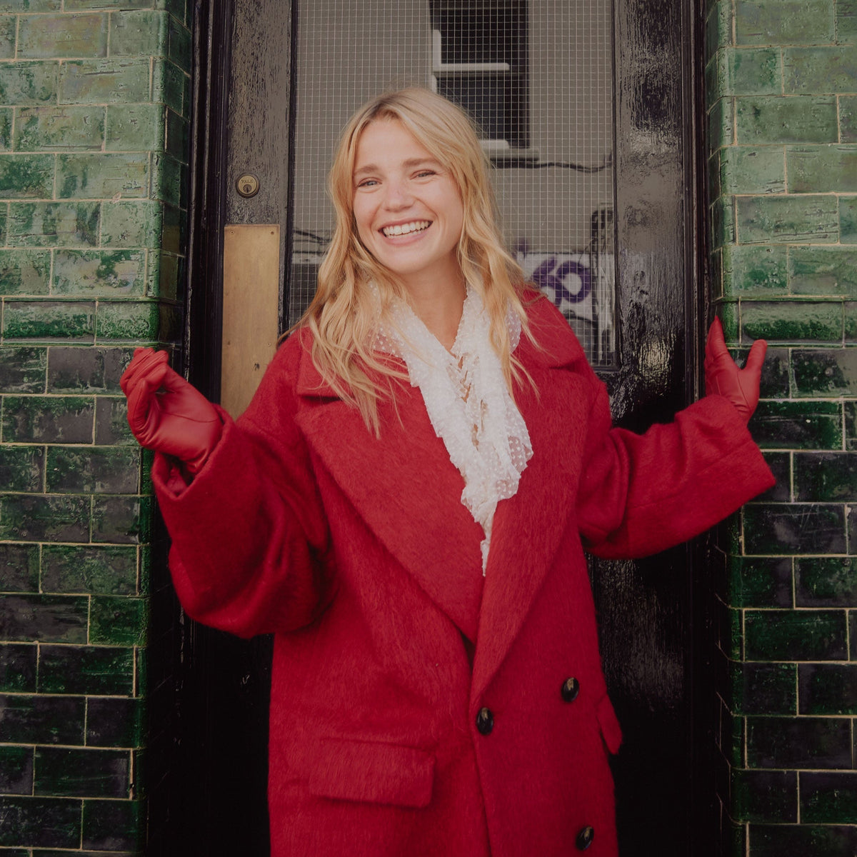 Woman stood in front of a deep green, brick wall wearing vibrant red leather gloves.