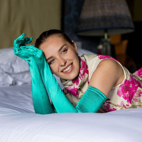 Woman wearing Women's long satin opera gloves holding her hands and on a bed in a pink dress. 