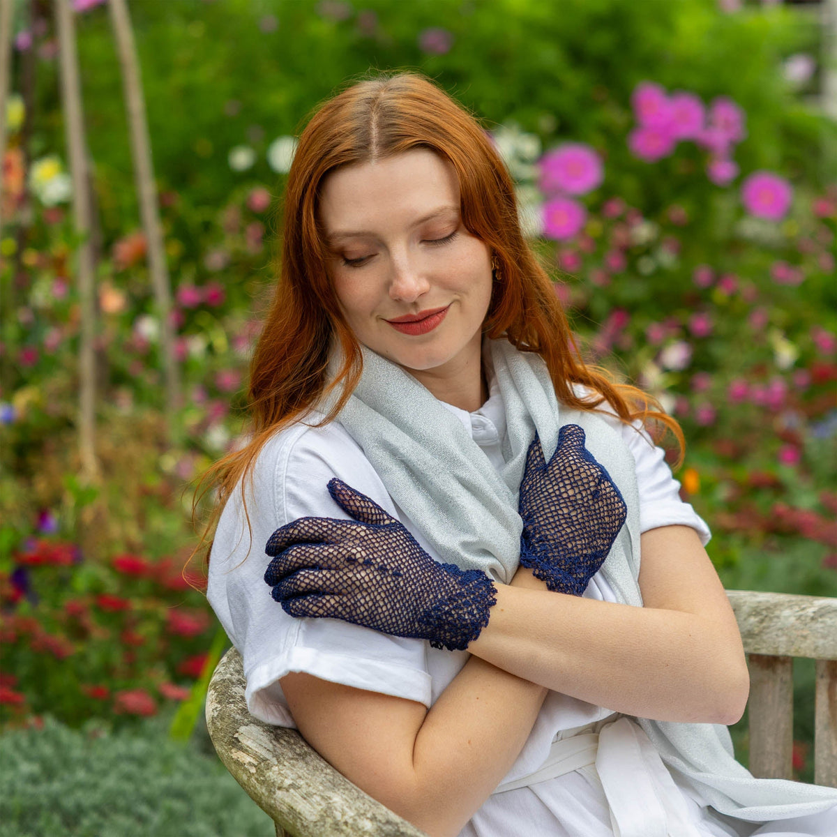 Woman wearing cotton crochet gloves in navy with silver scarf sitting on  bench in a garden 