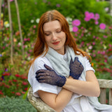 Woman wearing cotton crochet gloves in navy with silver scarf sitting on  bench in a garden 