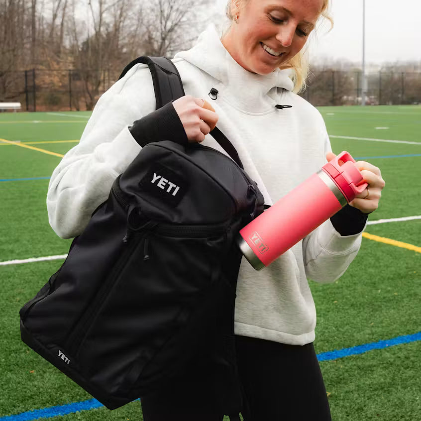 A woman on an athletic field carries a black YETI backpack and holds a pink YETI water bottle. The backpack features a white YETI logo on the front pocket.
