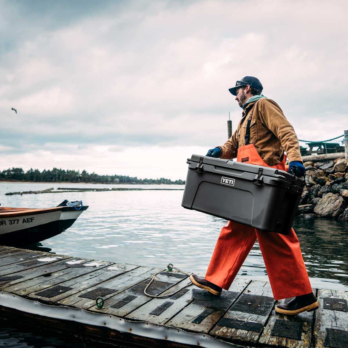 A man carrying a large YETI Charcoal  Tundra 45 cooler on a weathered wooden dock next to the water, wearing orange overalls, a brown jacket, and a blue cap under an overcast sky