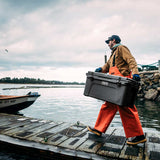 A man carrying a large YETI Charcoal  Tundra 45 cooler on a weathered wooden dock next to the water, wearing orange overalls, a brown jacket, and a blue cap under an overcast sky