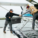 Two people on a seaplane at the waterline passing a large Charcoal Tundra 45 YETI cooler. One person is standing on the deck holding the cooler, while the other climbs onto the plane from the ramp holding a tan backpack.