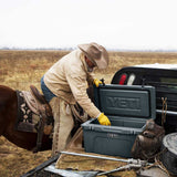 A person in western clothing leans over an open YETI cooler placed on the bed of a pickup truck in an open field. The cooler is dark gray with a YETI logo embossed on the lid and a smaller YETI label on the front.