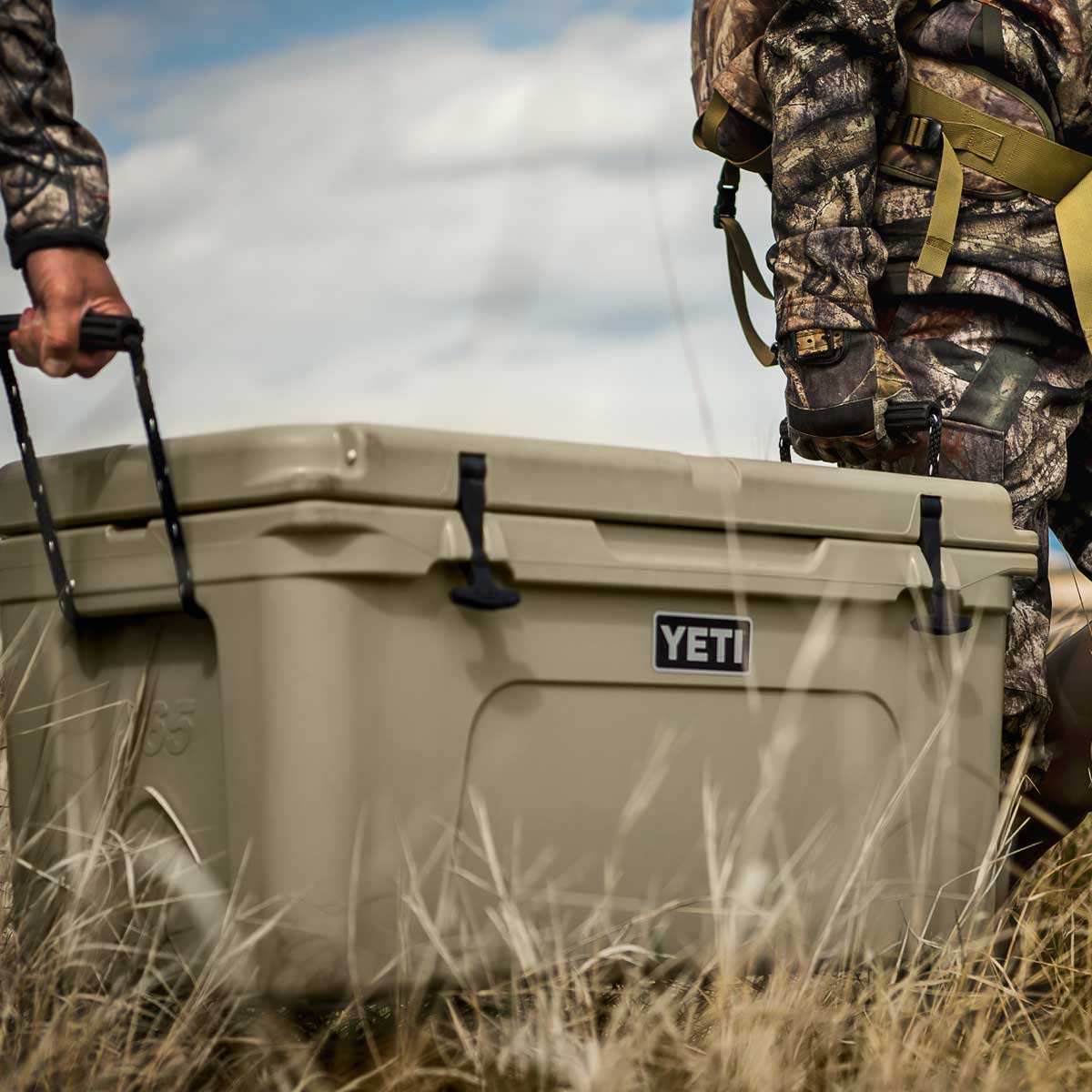 A YETI cooler in tan is being carried outdoors by two people in camouflage hunting gear, with dry grass and a cloudy sky in the background.