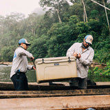 Two men in a wooden boat on a river handle a large Tan Tundra 65 YETI cooler. The background is a lush, green, misty forest.