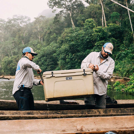 Two men in a wooden boat on a river handle a large Tan Tundra 65 YETI cooler. The background is a lush, green, misty forest.